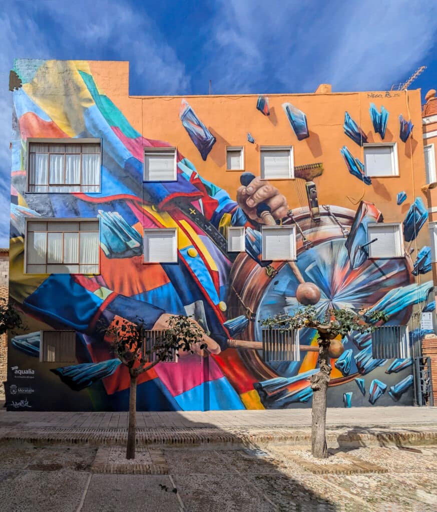 Un vibrante mural cubre un edificio y representa una colorida figura abstracta con traje que sostiene una peonza. Fragmentos de formas azules y rojas rodean la figura, mezclándose con las ventanas y la estructura de la fachada naranja.