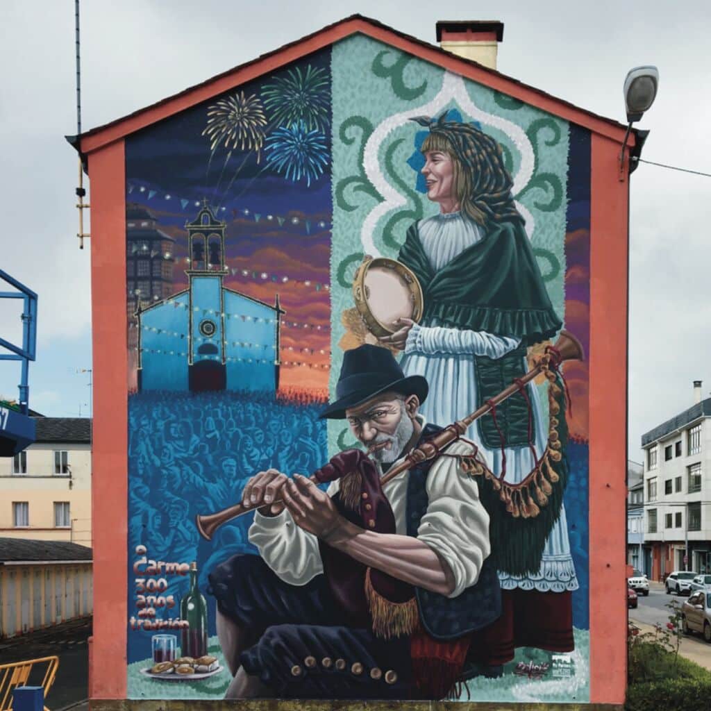 Colorido mural en un edificio que muestra a un hombre sentado tocando la gaita y a una mujer de pie con una pandereta; detrás de ellos hay una escena nocturna festiva con una iglesia, fuegos artificiales y una multitud bajo cuerdas de luces.