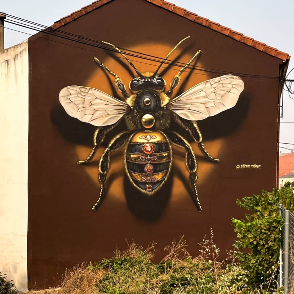 Un gran mural realista de una abeja con alas detalladas y brillantes detalles dorados está pintado en el lateral de un edificio marrón, rodeado de vegetación y líneas eléctricas.