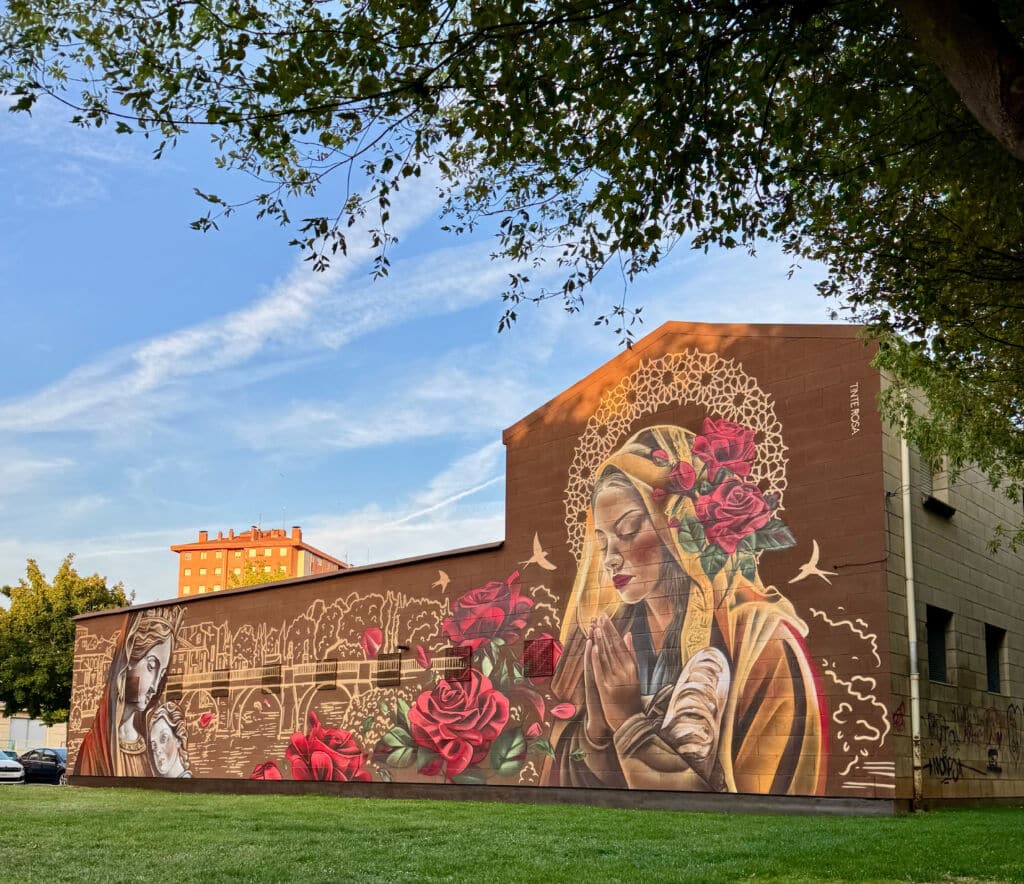 Un vibrante mural en un edificio muestra a dos mujeres, una con un bebé en brazos y otra con los ojos cerrados rezando, rodeadas de grandes rosas rojas, motivos dorados y pájaros, sobre un cielo azul y hierba verde.