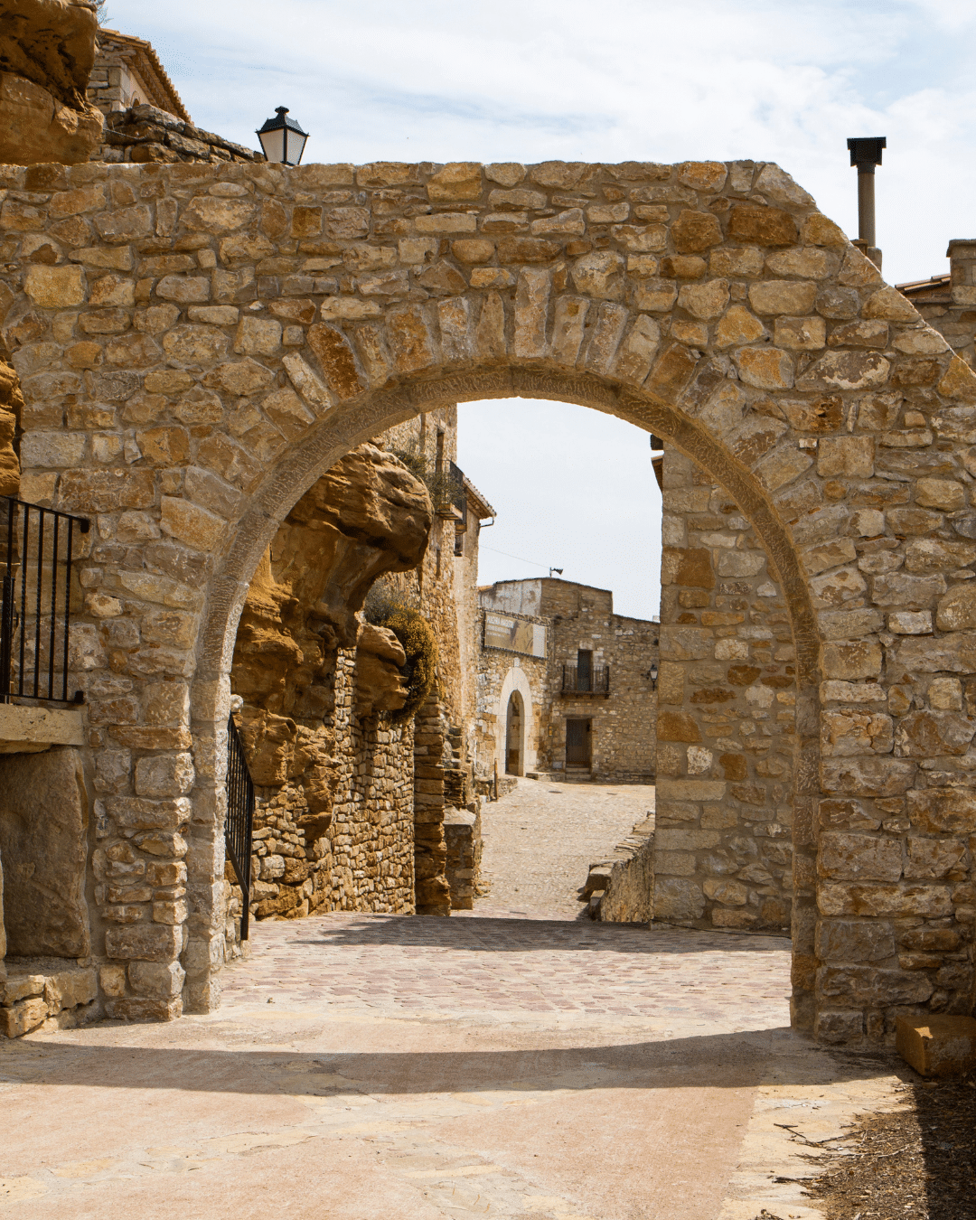 Un gran arco de piedra se alza en un tranquilo y rústico pueblo con viejos edificios de piedra y un estrecho camino, bajo un cielo parcialmente nublado.