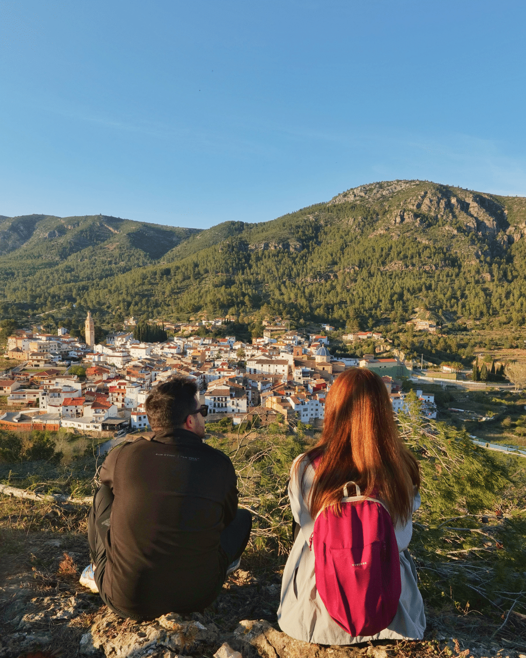 Dos personas están sentadas en una colina desde la que se domina una ciudad con edificios de tejados rojos, rodeada de verdes colinas y montañas bajo un cielo azul despejado. Una persona lleva una chaqueta negra y la otra tiene el pelo largo y una mochila roja.