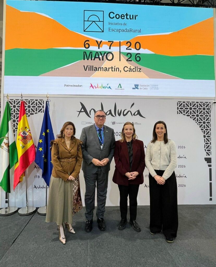 Cuatro personas de pie delante de un expositor que promociona un evento de turismo rural en Villamartín, Cádiz, con banderas y una gran pantalla que muestra detalles del evento y logotipos detrás de ellos.