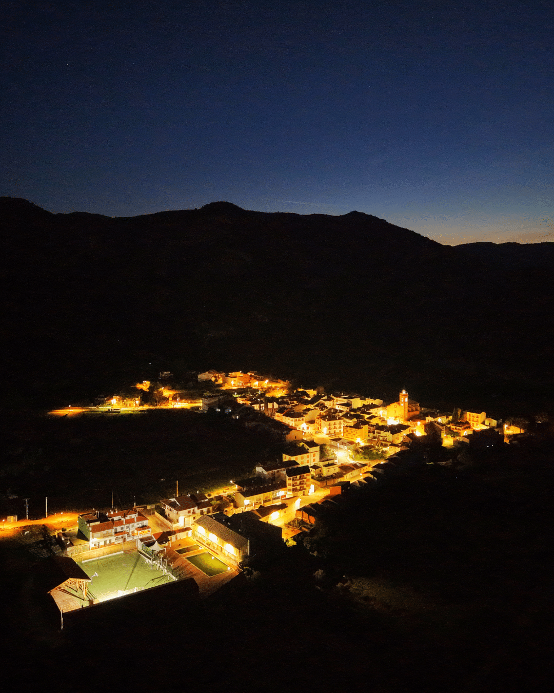 Una pequeña ciudad enclavada en un valle se ilumina de noche con farolas, rodeada de montañas oscuras bajo un cielo despejado y estrellado.