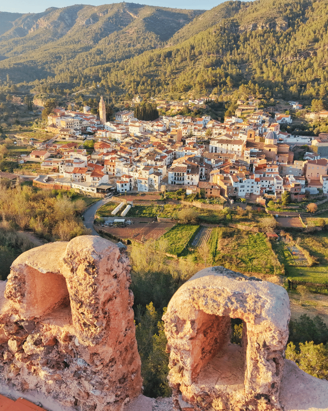 Vista panorámica de una pequeña ciudad con tejados de tejas rojas enclavada entre verdes colinas, vista desde un antiguo muro de piedra en primer plano. La luz del sol proyecta tonos cálidos sobre el paisaje, resaltando la torre de la iglesia y los edificios circundantes.