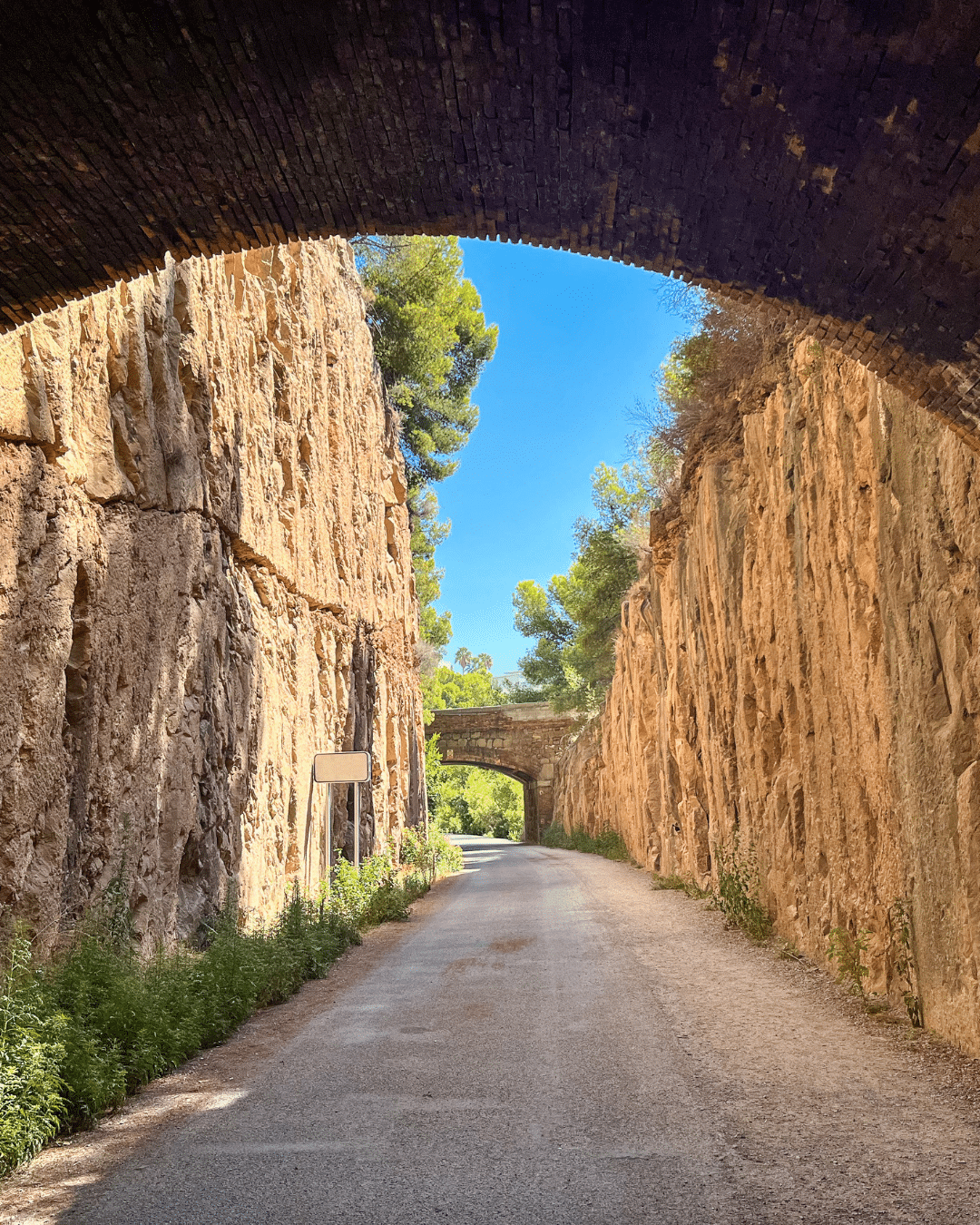 Una carretera asfaltada pasa entre altos acantilados rocosos, con dos puentes de piedra sobre ellos, rodeados de vegetación y árboles bajo un cielo azul brillante.