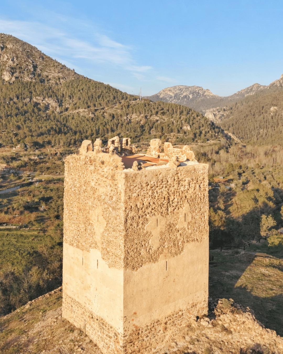 Una alta torre cuadrada de piedra con la parte superior parcialmente derruida se alza en la ladera de una colina, rodeada de verde vegetación y montañas bajo un cielo azul despejado.