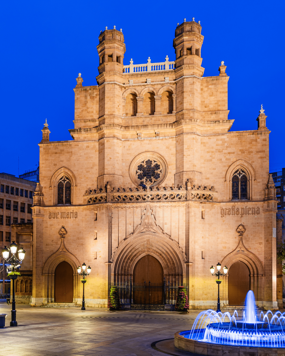 Una iglesia de piedra de estilo gótico con portadas arqueadas, torres y un rosetón, iluminada por la noche. En primer plano, una pequeña fuente con agua iluminada en azul en una plaza de la ciudad.