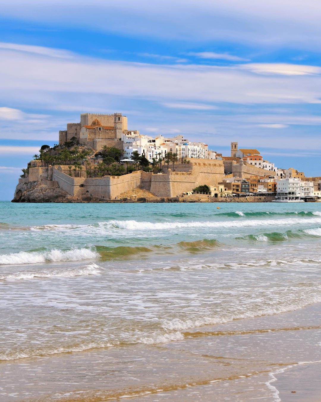 Un castillo histórico y edificios blancos se asientan sobre una colina rocosa que domina el mar, con suaves olas que bañan una playa de arena en primer plano bajo un cielo parcialmente nublado.