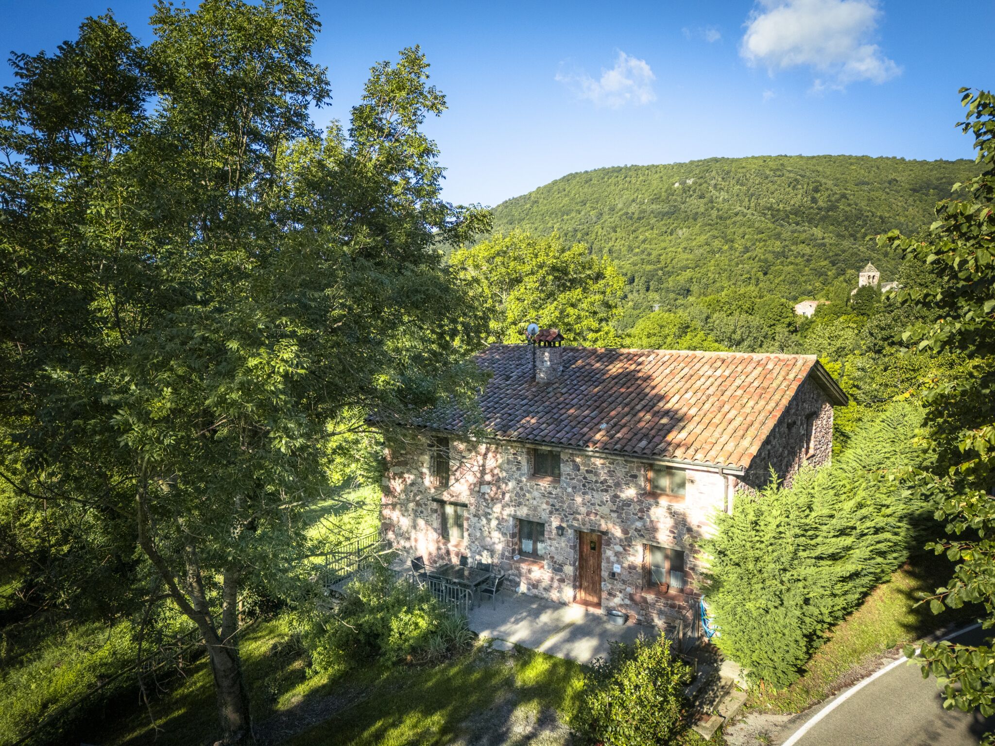 Una rústica casa de piedra con tejado de tejas está rodeada de frondosos árboles verdes y colinas bajo un cielo azul brillante con algunas nubes. Junto a la casa discurre una pequeña carretera, y al fondo se eleva una colina cubierta por un denso bosque.