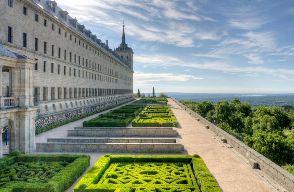 Un gran edificio de piedra con hileras de ventanas da a unos setos verdes de formas geométricas y a un cuidado jardín, con un camino al lado y un pintoresco paisaje de árboles y cielo al fondo.