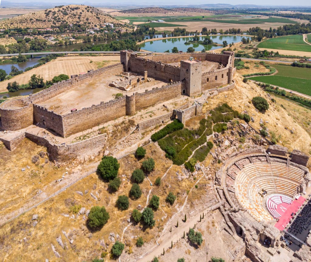 Vista aérea de una antigua fortaleza de piedra en lo alto de una colina con un anfiteatro circular cerca, rodeada de tierras de labranza, ríos y colinas onduladas bajo un cielo despejado.