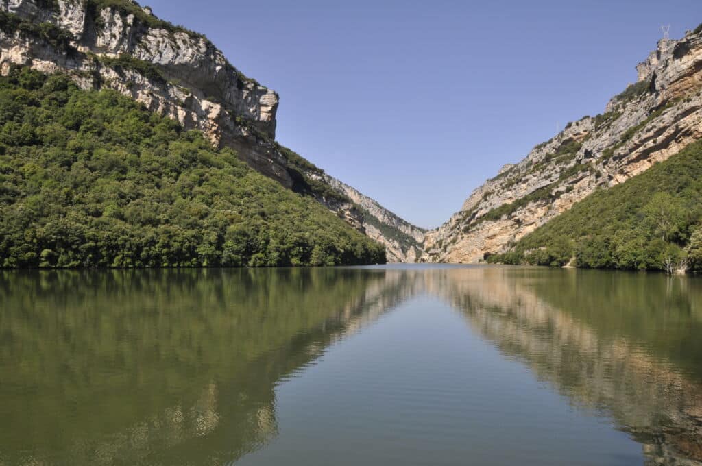 Un río tranquilo fluye entre dos escarpadas laderas montañosas boscosas bajo un cielo azul despejado, con el paisaje reflejado en el agua.