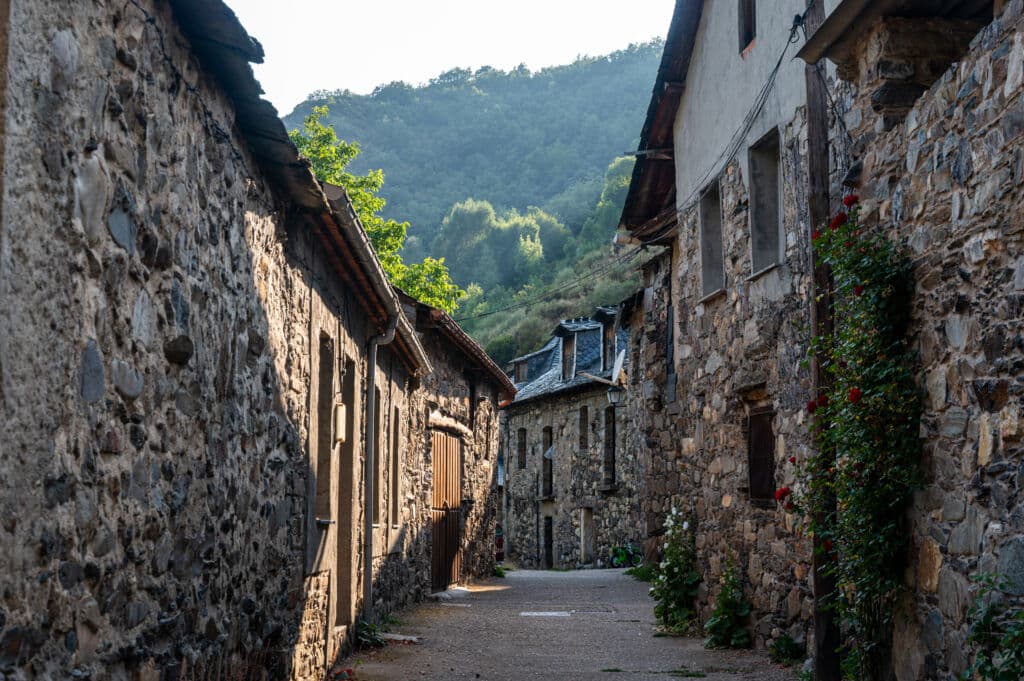 Una estrecha calle empedrada bordeada de viejas casas de piedra y flores rojas, situada en un tranquilo pueblo de montaña con verdes colinas al fondo.