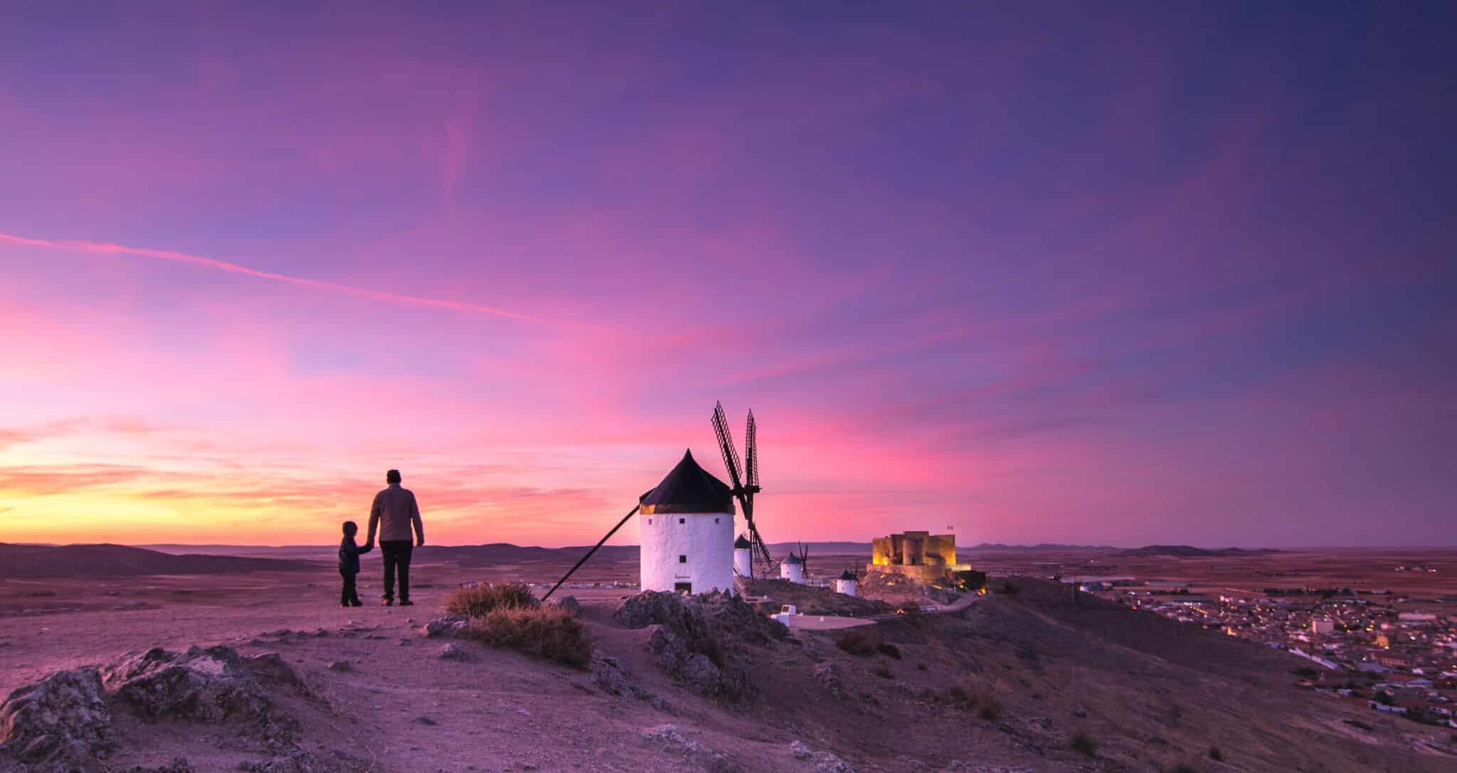 Una persona y un niño cerca de unos molinos de viento tradicionales al atardecer, contemplando un vasto paisaje con un pueblo a lo lejos bajo un vibrante cielo púrpura y rosa.