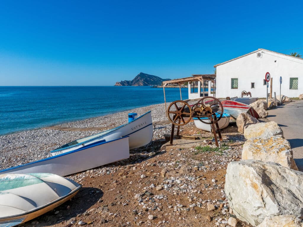 Varias barcas pequeñas descansan en una playa rocosa junto a un edificio blanco, con el mar azul en calma y una montaña lejana bajo un cielo claro y brillante. Unas ruedas de metal oxidado descansan cerca de las barcas en la orilla.