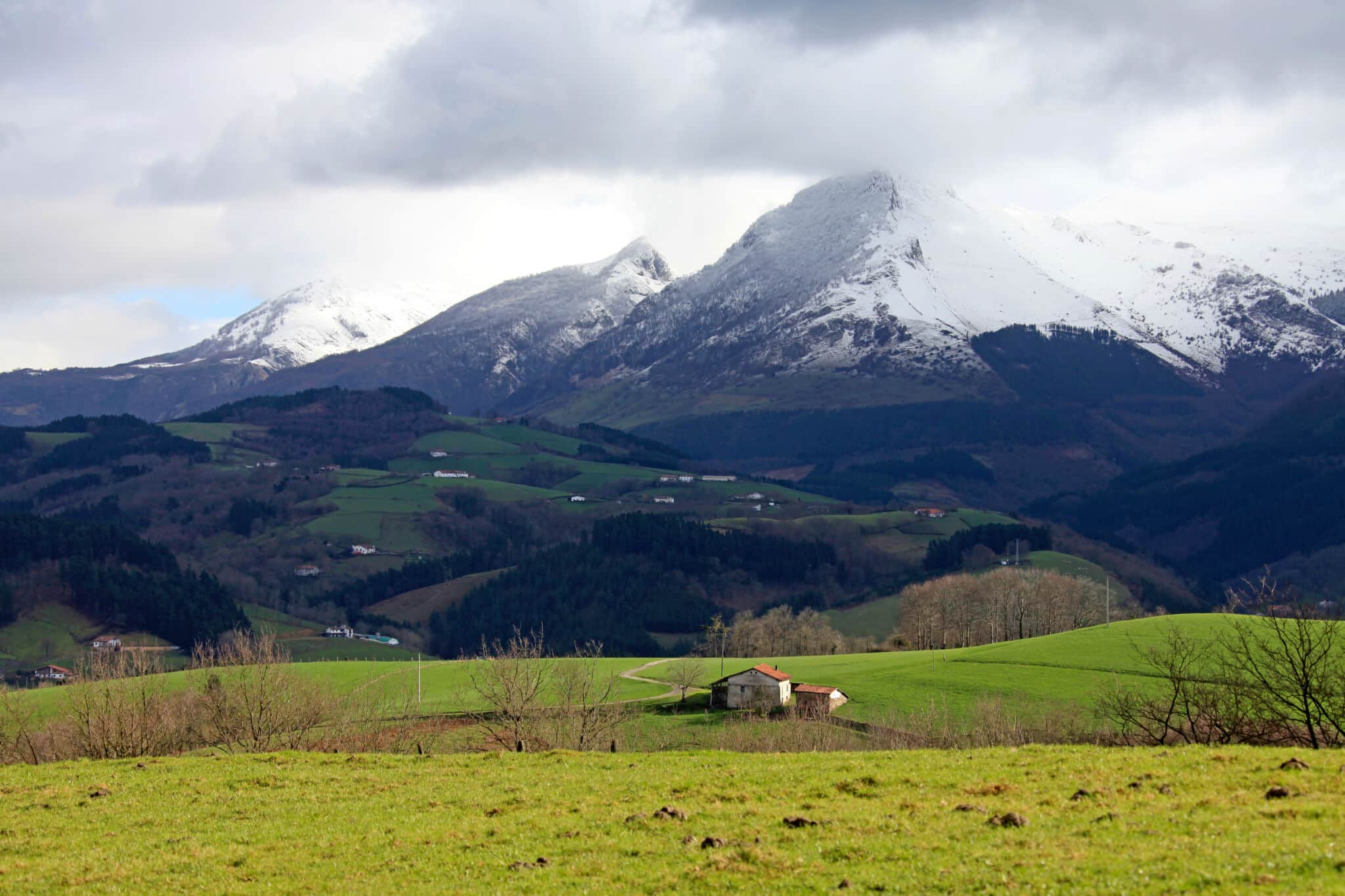 Una pequeña casa se asienta sobre una verde colina cubierta de hierba, rodeada de árboles, con montañas nevadas y nubes dispersas al fondo.