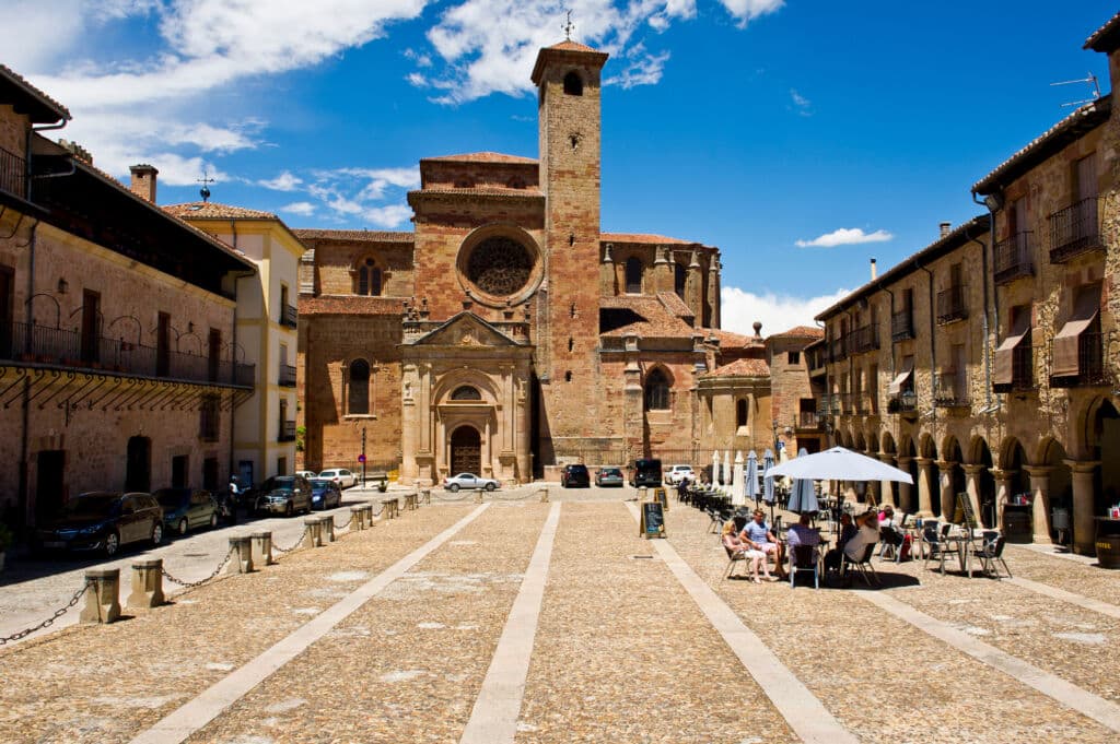 Plaza soleada con un camino empedrado que conduce a una gran iglesia histórica de piedra. La gente está sentada bajo sombrillas en un café al aire libre a la derecha, con edificios antiguos y coches aparcados bordeando la plaza. Cielo azul.