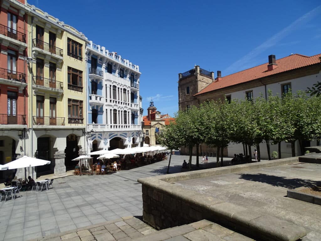 Plaza soleada con cafeterías al aire libre bajo sombrillas blancas, rodeada de coloridos edificios históricos, frondosos árboles y un cielo azul despejado. Unos escalones de piedra conducen a la plaza.
