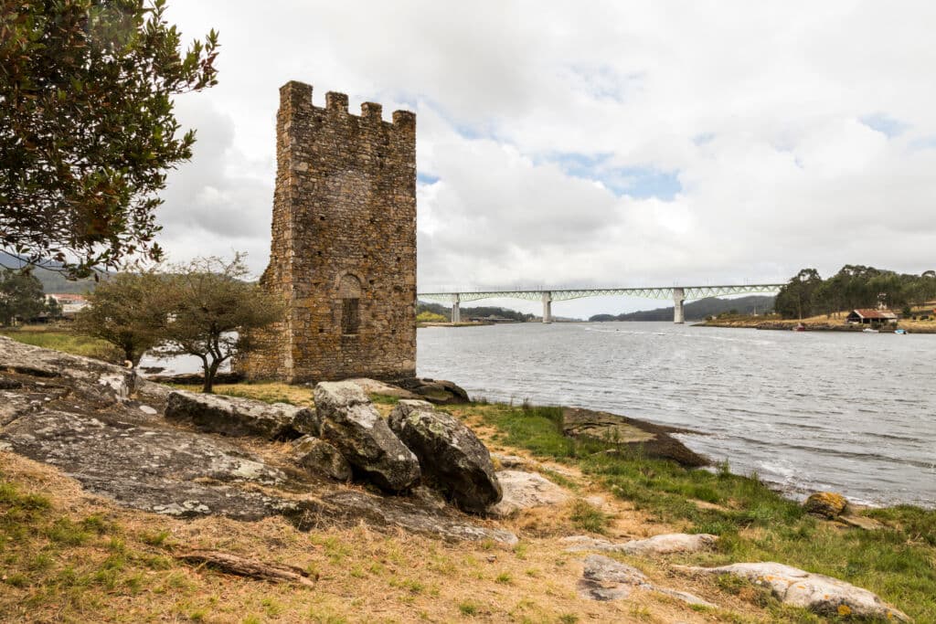 Una pequeña torre medieval de piedra se alza junto a la orilla de un río con suelo rocoso y hierba, un árbol cerca y un puente moderno que cruza el río al fondo, bajo un cielo nublado.