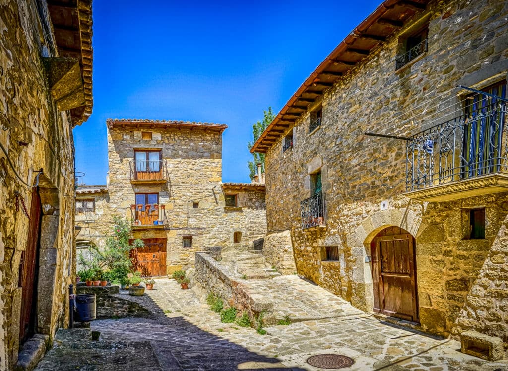 Una estrecha calle adoquinada bordeada de rústicos edificios de piedra, balcones de madera y portales arqueados bajo un cielo azul brillante en un pueblo pintoresco y soleado.