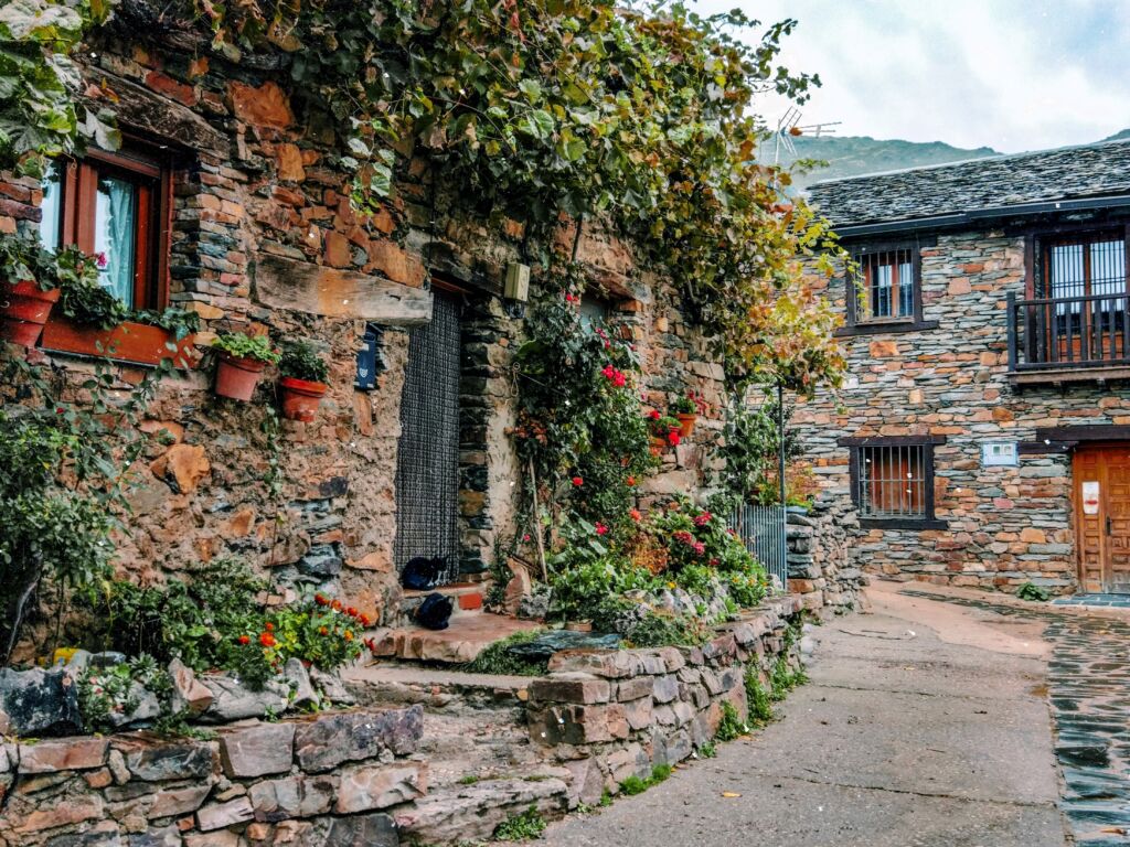 Una encantadora casa de piedra con plantas trepadoras y macetas en sus paredes se alza junto a un estrecho y sinuoso camino en un pueblo rústico. Al fondo se ve otro edificio de piedra bajo un cielo nublado.