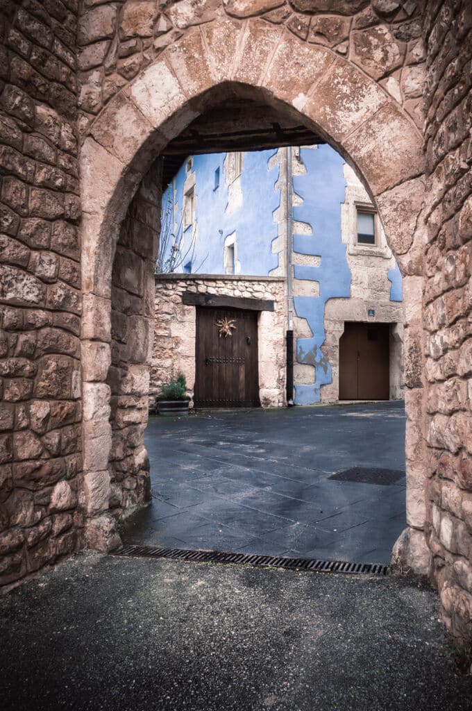 Un arco de piedra enmarca la vista de un patio empedrado con un edificio azul brillante, puertas de madera y pequeñas ventanas en un pintoresco entorno de pueblo europeo.