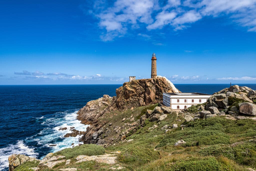 Un faro se alza en un acantilado rocoso sobre el océano, con las olas rompiendo contra la orilla. En primer plano, un edificio blanco entre arbustos verdes bajo un cielo azul brillante con nubes dispersas.