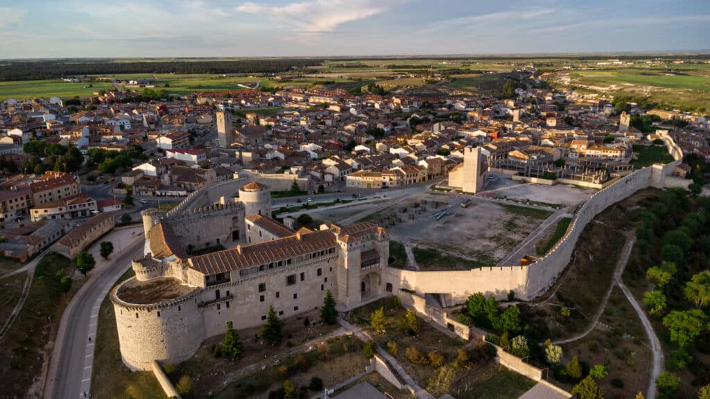 Vista aérea de un gran castillo histórico de piedra con torres y murallas fortificadas, rodeado por una pequeña ciudad con tejados de tejas rojas y campos verdes abiertos al fondo bajo un cielo parcialmente nublado.