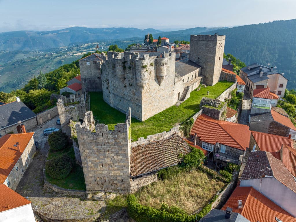 Vista aérea de un castillo medieval de piedra con torres, rodeado de casas de tejados rojos y verdes colinas al fondo bajo un cielo despejado.