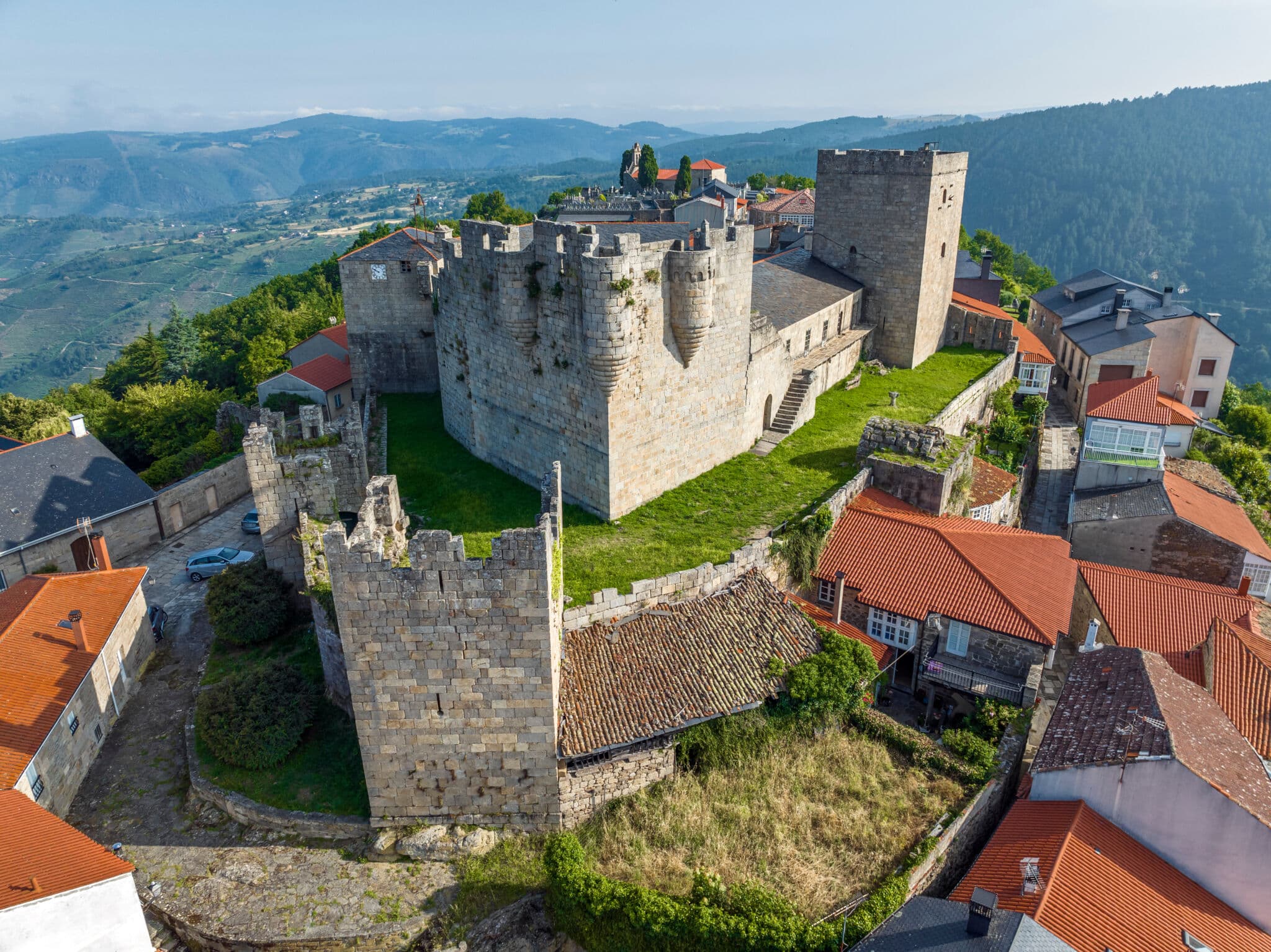 Vista aérea de un castillo medieval de piedra con torres, rodeado de casas de tejados rojos y verdes colinas al fondo bajo un cielo despejado.
