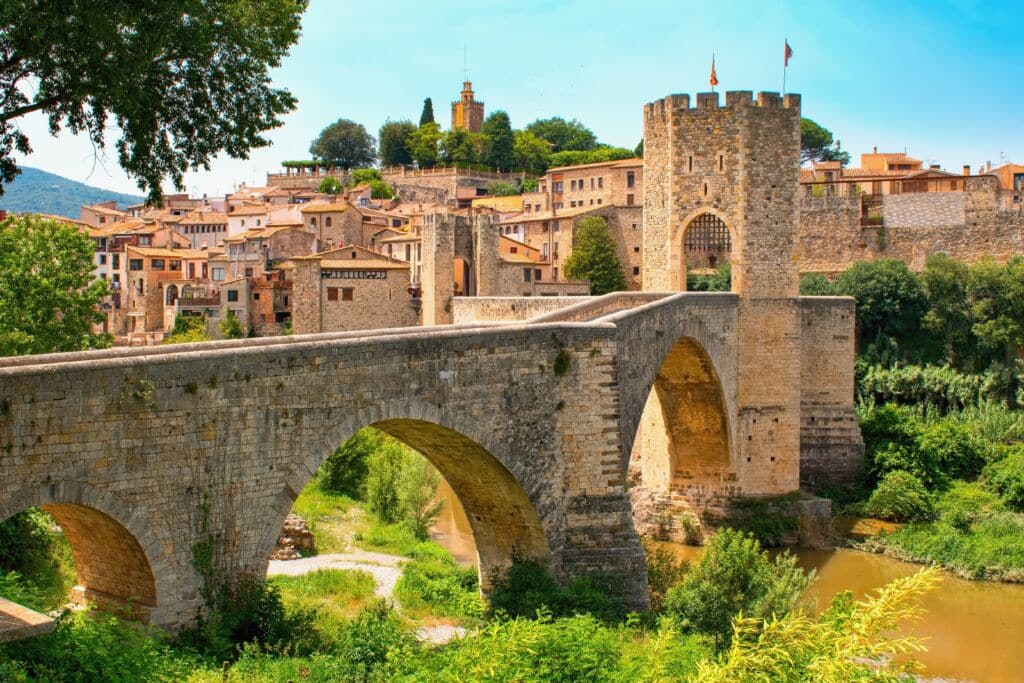 Un puente medieval de piedra con arcos y una puerta fortificada cruza un río y conduce a un pueblo histórico con edificios rústicos, árboles y una torre al fondo, bajo un cielo claro y brillante.