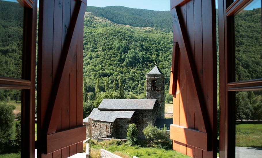 Vista a través de contraventanas de madera abiertas de una iglesia de piedra con campanario, rodeada de exuberantes colinas verdes y densos bosques bajo un cielo despejado.