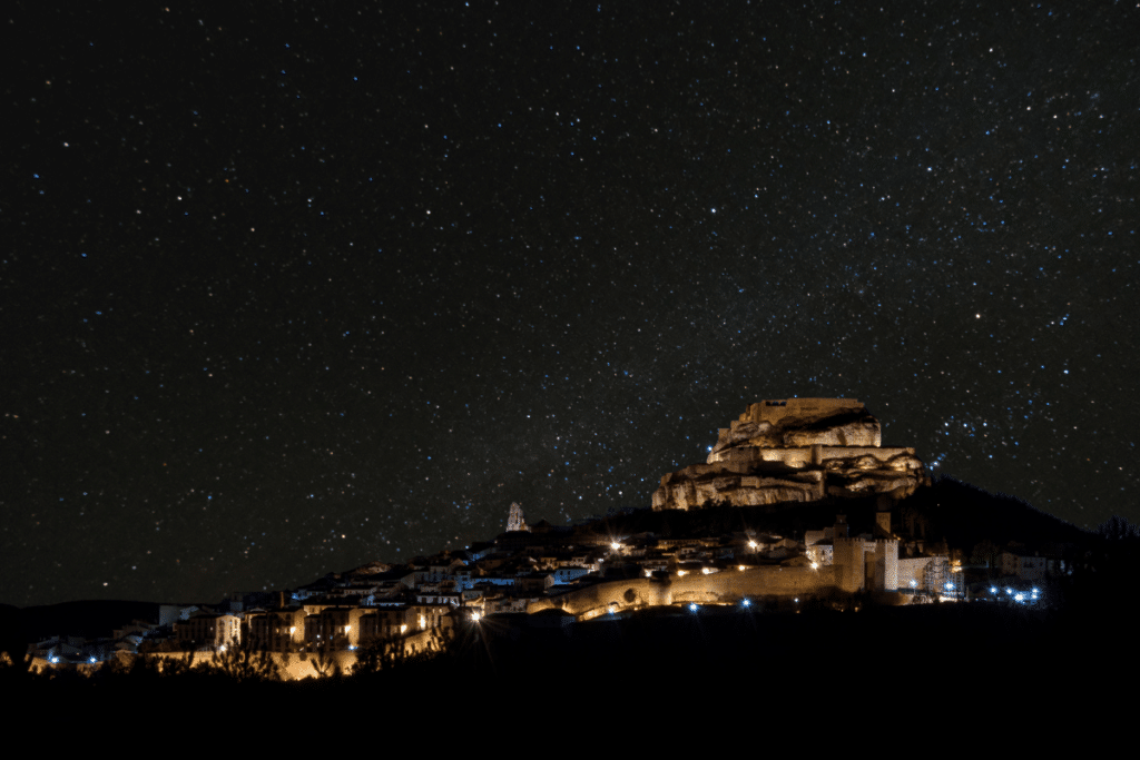 Una ciudad en lo alto de una colina con edificios iluminados y un castillo se ve de noche bajo un cielo despejado y lleno de estrellas. La escena está rodeada de oscuridad, lo que resalta las cálidas luces de la ciudad y la inmensidad del cielo nocturno.