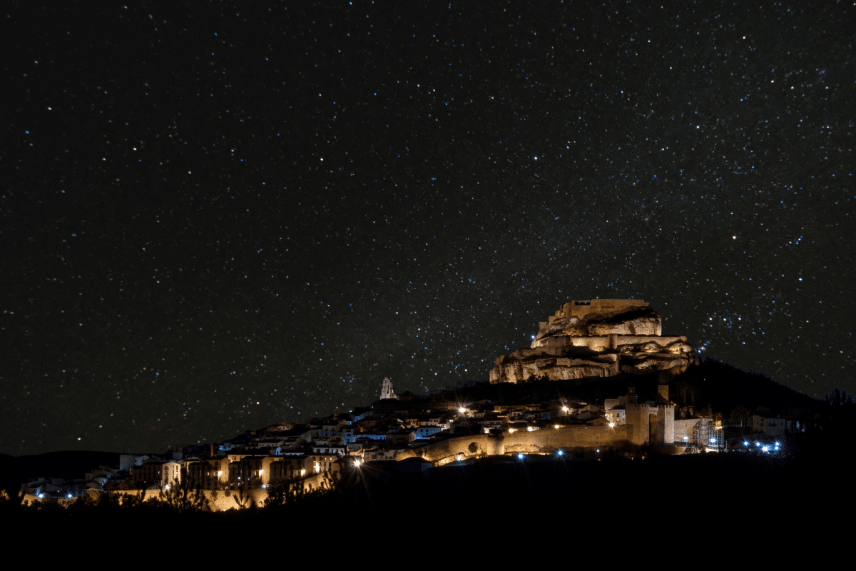 Una ciudad en lo alto de una colina con edificios iluminados y un castillo se ve de noche bajo un cielo despejado y lleno de estrellas. La escena está rodeada de oscuridad, lo que resalta las cálidas luces de la ciudad y la inmensidad del cielo nocturno.