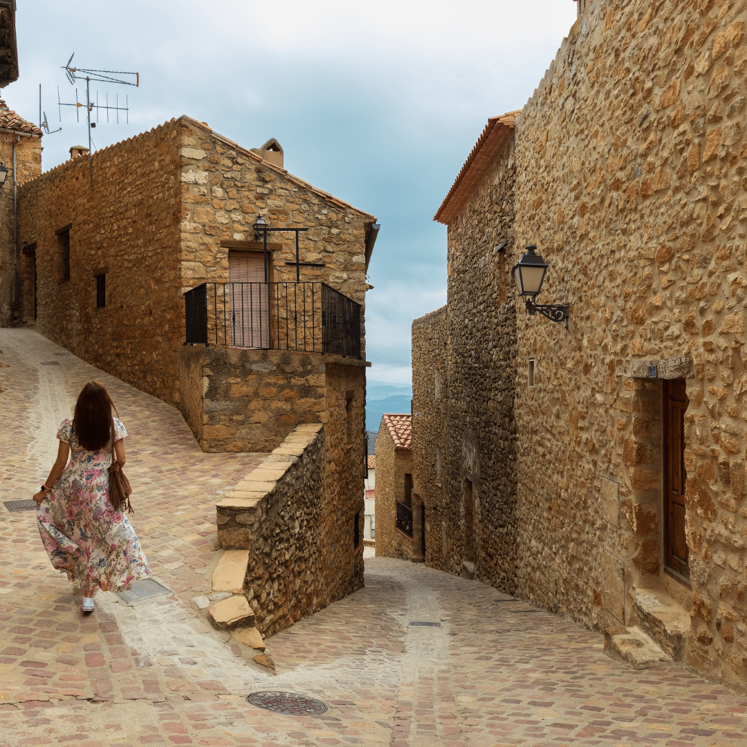 Una mujer con un vestido de flores camina por una estrecha y sinuosa calle empedrada bordeada de viejos edificios de piedra bajo un cielo nublado.