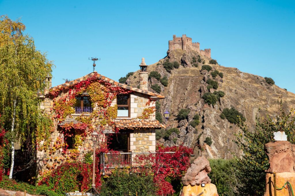 Una casa de piedra cubierta de hiedra otoñal se alza en primer plano, con una colina rocosa y un castillo medieval encaramado en la cima al fondo, todo ello bajo un cielo azul despejado.