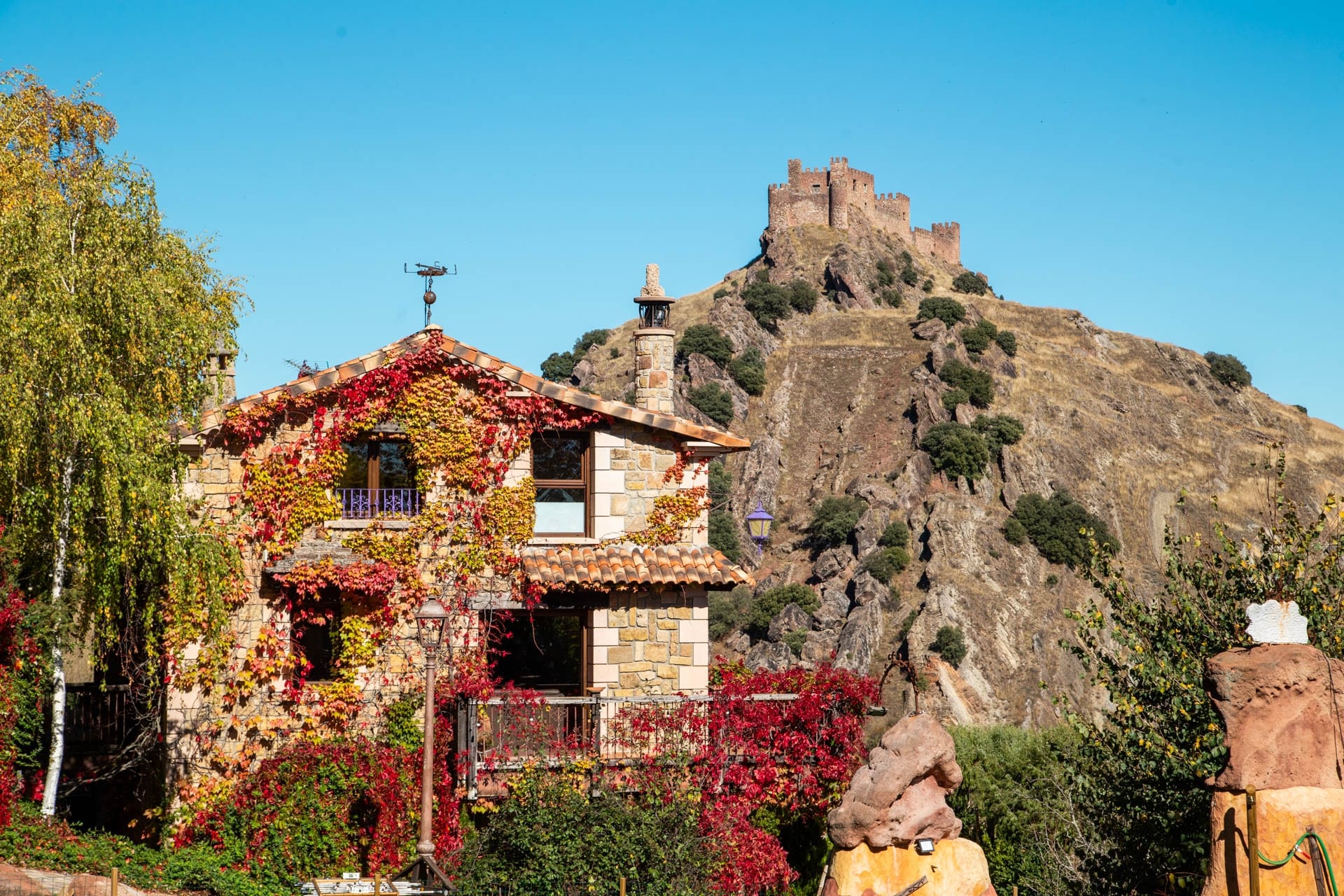 Una casa de piedra cubierta de hiedra otoñal se alza en primer plano, con una colina rocosa y un castillo medieval encaramado en la cima al fondo, todo ello bajo un cielo azul despejado.