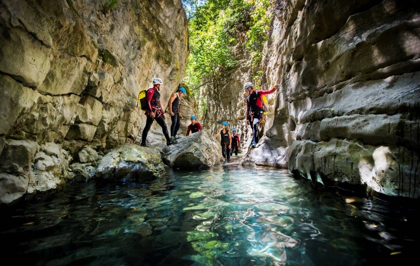 Un grupo de personas con casco y traje de neopreno exploran un estrecho cañón rocoso de aguas claras y poco profundas, rodeado de altos muros de piedra y vegetación.