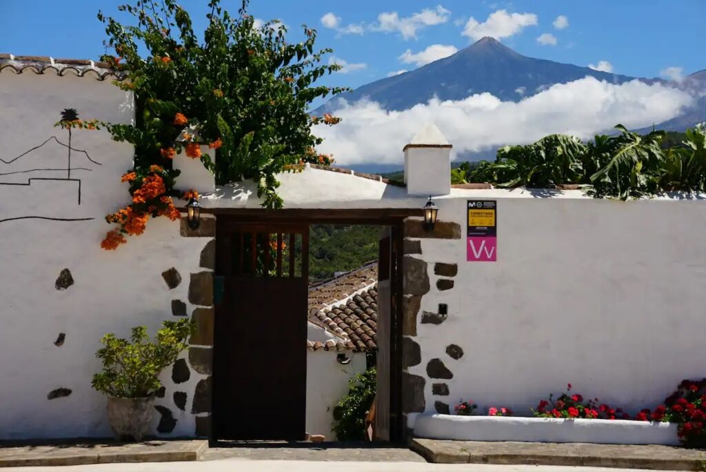 Un patio de paredes blancas con una puerta de madera, flores trepadoras y un paisaje montañoso de fondo. En el interior se ve un tejado de tejas y flores vibrantes, con un pico nevado y un cielo azul más allá.