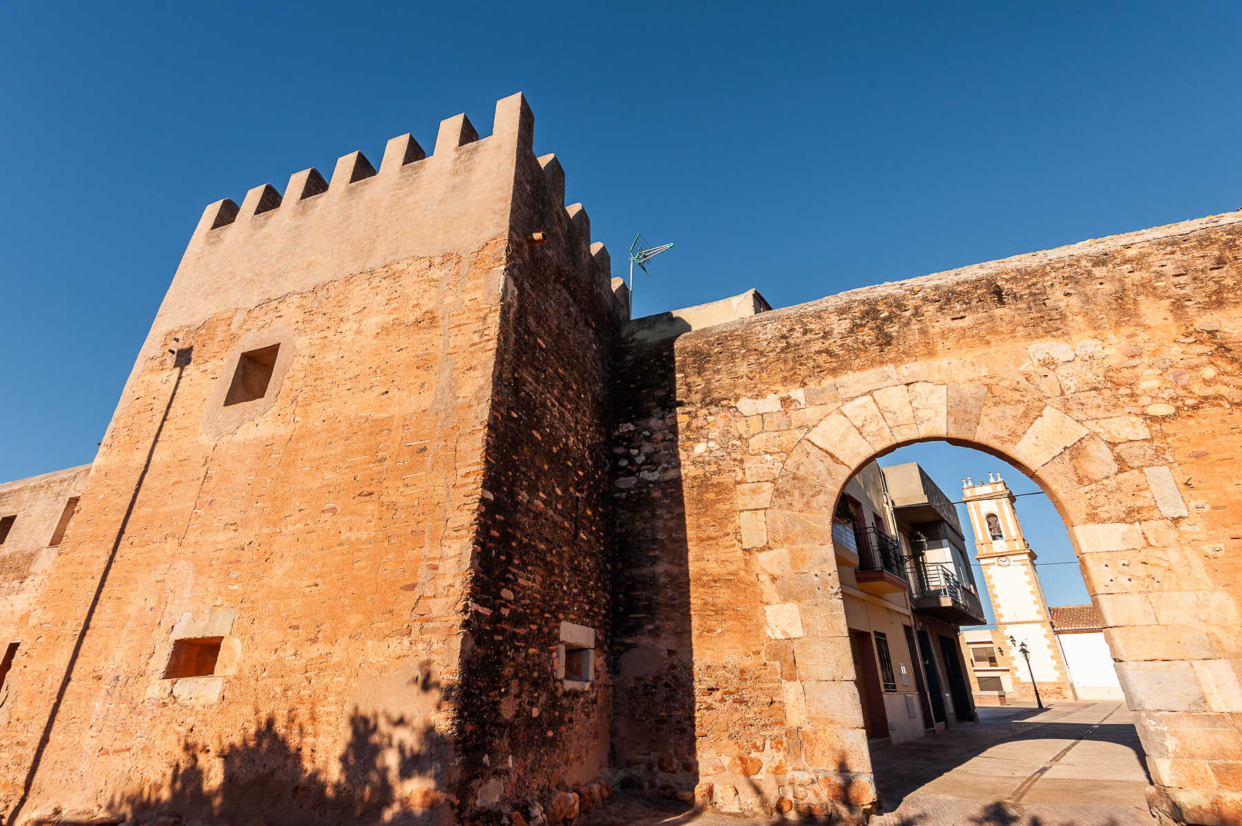 Un histórico muro de piedra con almenas y una puerta arqueada se alza bajo un cielo azul despejado, con una torre del reloj y edificios visibles a través del arco.
