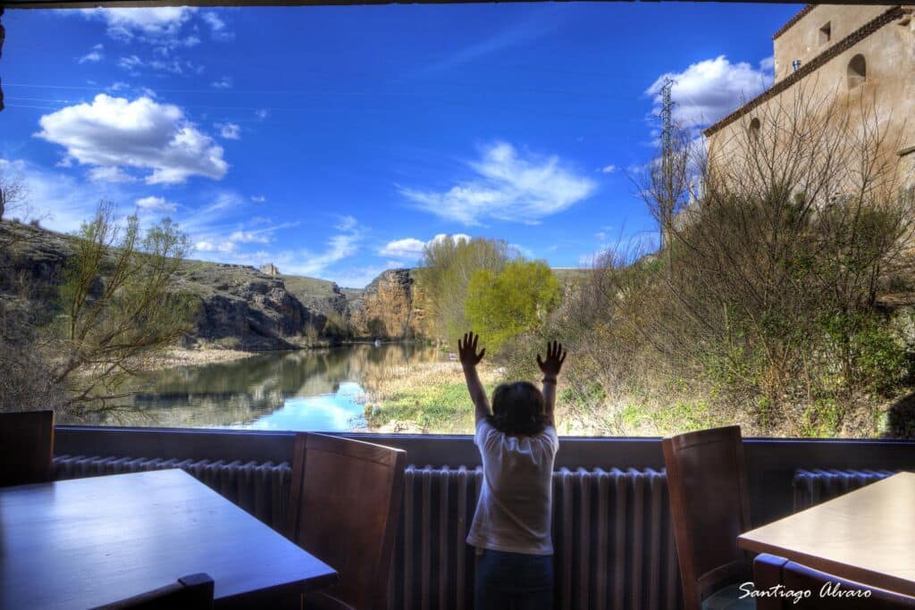 Un niño está de pie junto a una gran ventana, levantando ambas manos hacia una vista panorámica de un río, árboles y un cielo azul, rodeado de mesas y sillas vacías.