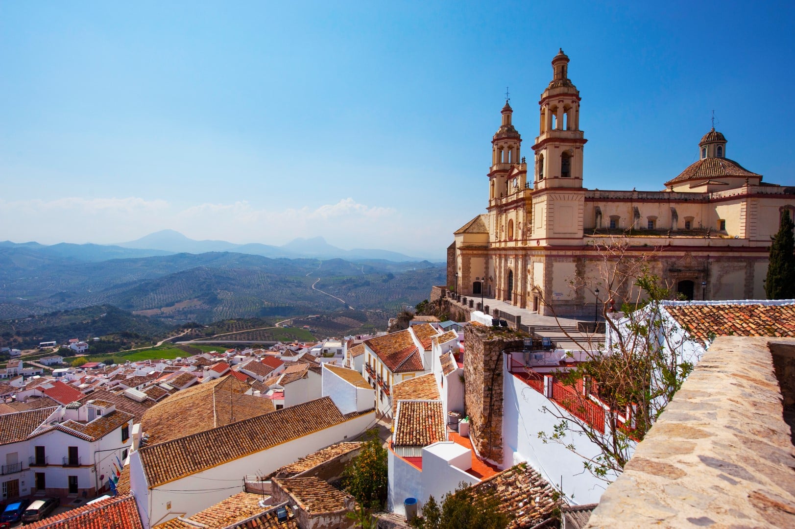 Una gran catedral con dos torres domina una ciudad en la ladera de una colina con edificios encalados y tejados de tejas rojas, con un telón de fondo de colinas onduladas bajo un cielo azul despejado.