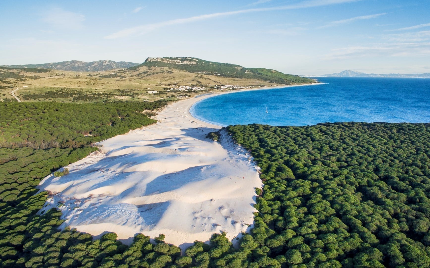 Vista aérea de una duna de arena bordeada por un bosque verde, una costa curva y un mar azul, con colinas lejanas y un pequeño asentamiento bajo un cielo despejado.