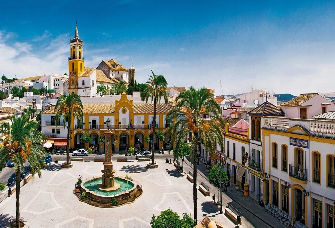 Una animada plaza con una fuente central, palmeras, edificios de colores y una iglesia con campanario bajo un cielo azul despejado. Hay gente paseando y coches aparcados a lo largo de la calle.