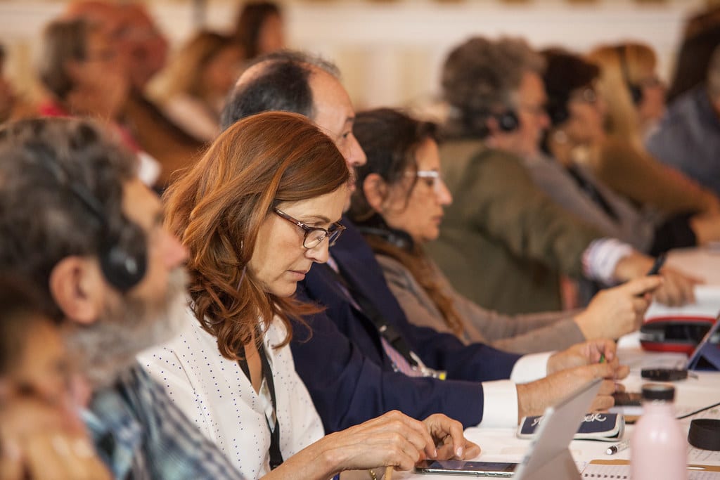 Un grupo de personas sentadas muy juntas en una conferencia o reunión de coetur, con los auriculares puestos y concentradas en sus ordenadores portátiles, notebooks o dispositivos móviles. El ambiente parece atento y comprometido.