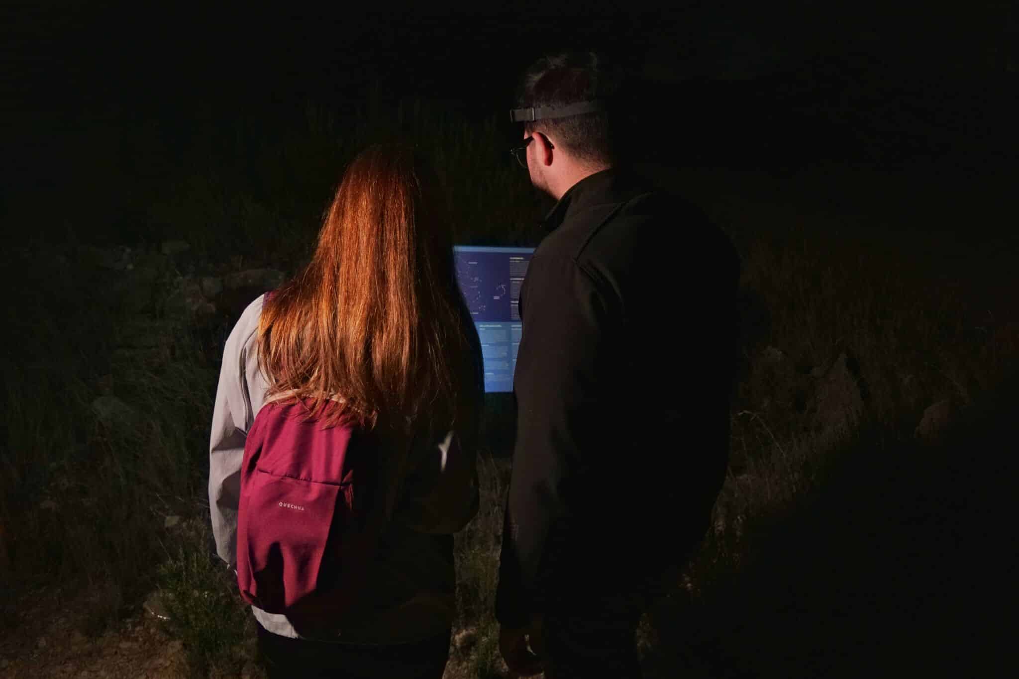 Dos personas, una de ellas con una mochila roja, están de pie al aire libre, en la oscuridad, mirando la pantalla iluminada de un ordenador portátil. Están de espaldas a la cámara y la escena está débilmente iluminada.