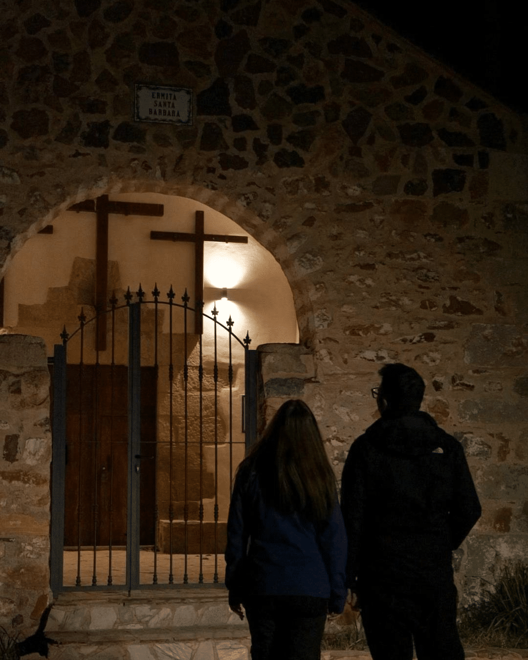 Por la noche, una pareja permanece de pie, uno al lado del otro, frente a un edificio de piedra con una entrada arqueada y una cruz iluminada por una luz cálida sobre la puerta.