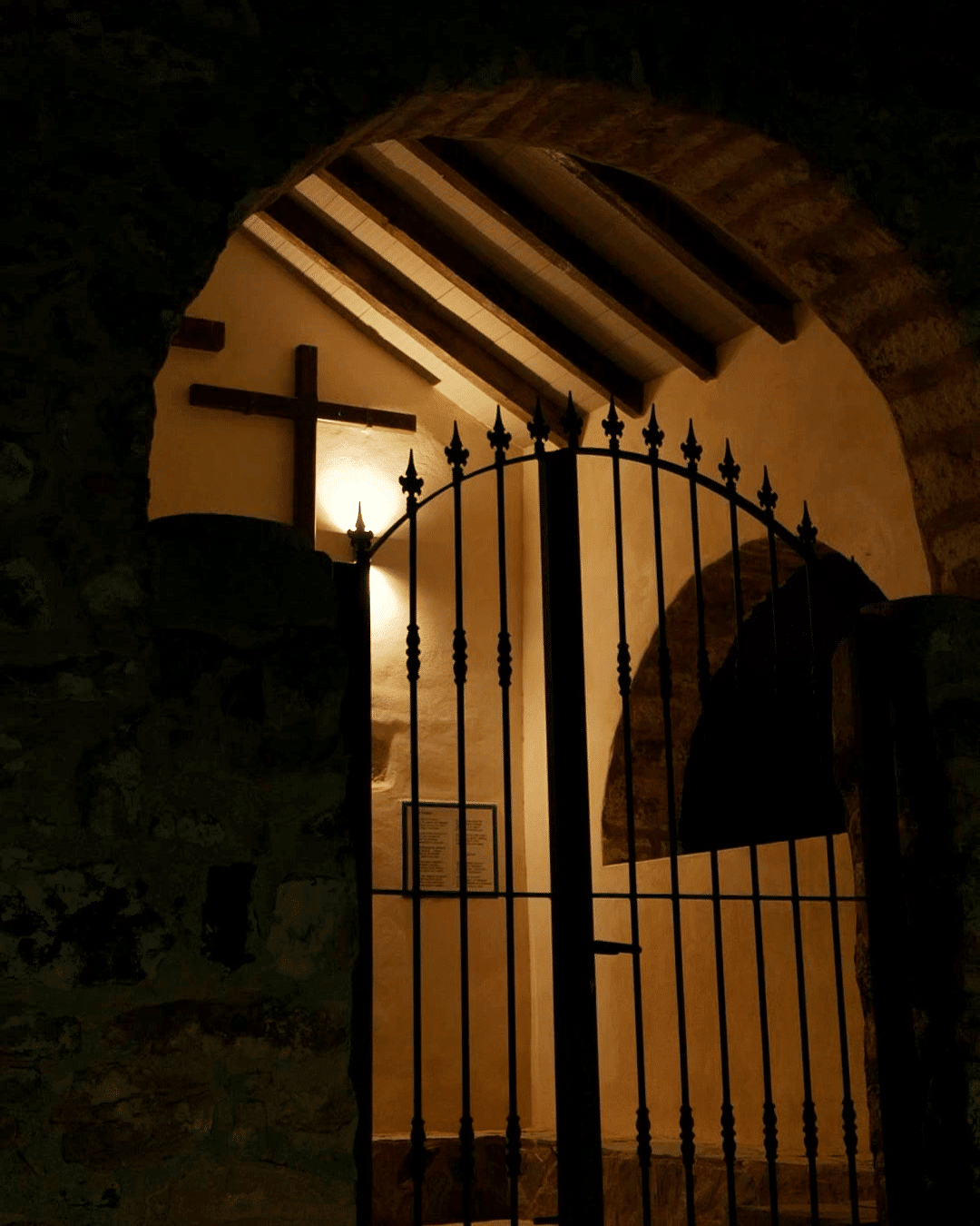 Un portón de hierro se alza de noche bajo un arco de piedra, iluminado por una luz cálida. En la pared interior hay una cruz de madera, y al fondo se ven vigas de madera en el techo y una placa enmarcada.