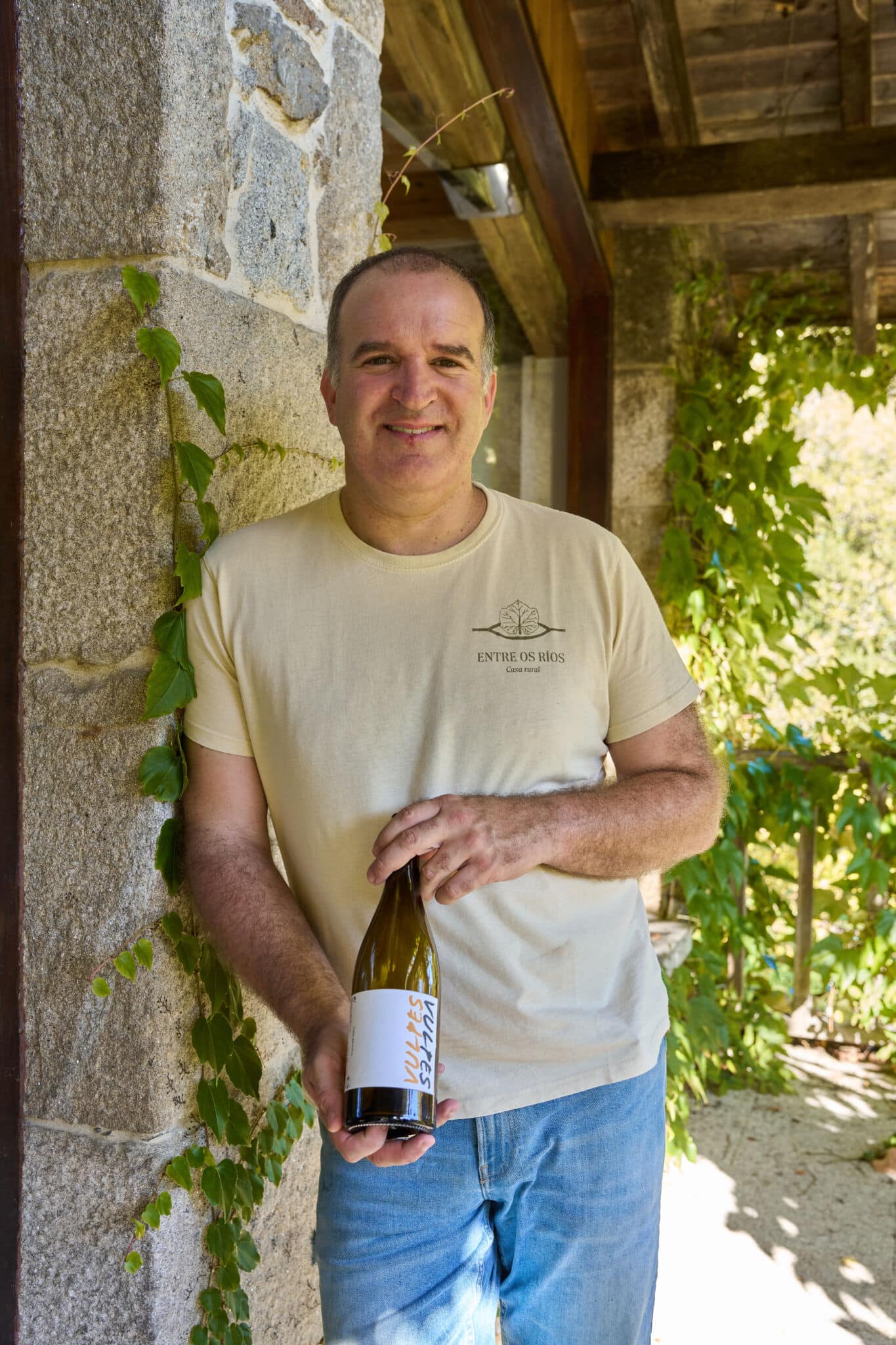 Un hombre con camiseta beige y vaqueros azules está de pie contra un muro de piedra cubierto de enredaderas verdes, sonriendo y sosteniendo una botella de vino al aire libre en un día soleado.